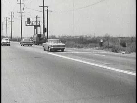 Cars driving along a two-lane highway surrounded by fields, vintage educational film.