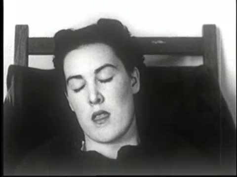 Woman resting in chair with eyes closed, head nodding slightly, static medium shot.