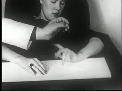 Hypnotized woman at table; a hand pricks her right hand while she remains still.