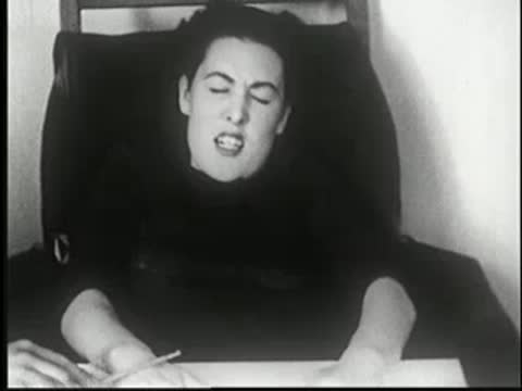 Hypnotized woman wincing in pain at table as an object touches her arm.