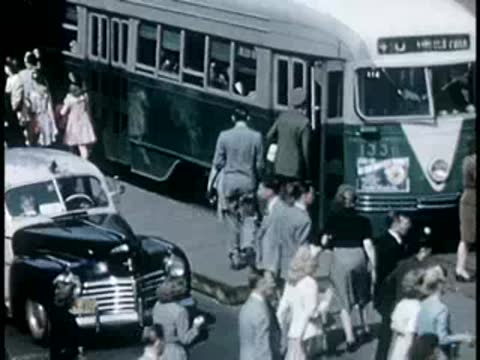 People boarding a green streetcar at a city stop, mid-20th-century clothing.