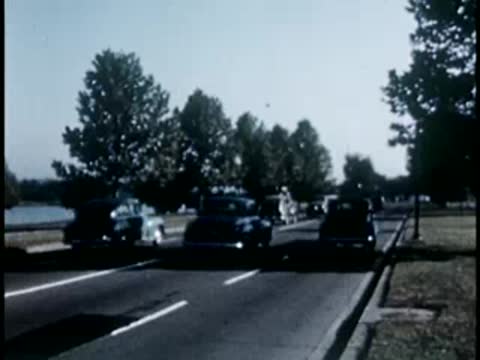 Car driving fast along a scenic lakeside road lined with trees, 1940s.