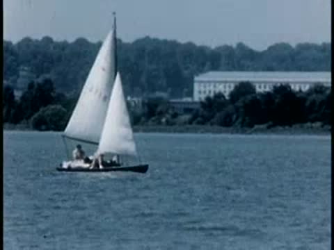 Small sailboat sailing across a calm body of water near a shore, 1940s.