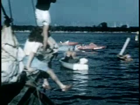 People on a sailboat watching speedboats racing across the water, mid-20th century.