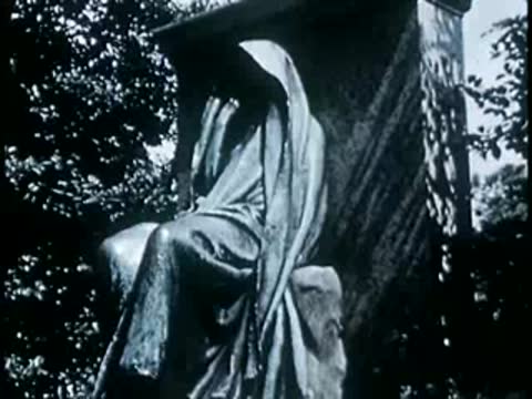 Statue of a hooded figure seated against a stone background