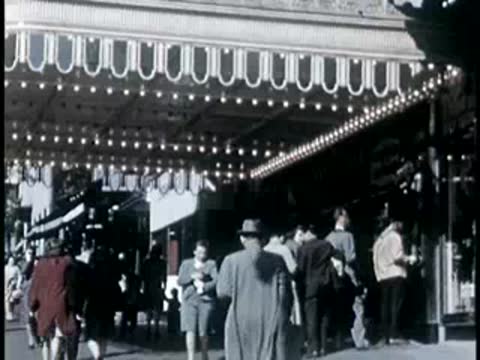 People walking past a lit marquee entrance in a city street, 1940s style.