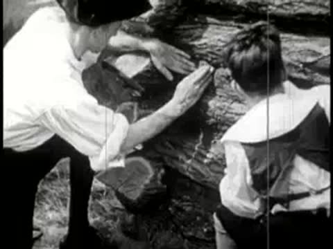 Two pioneers smoothing wet mud between stacked log walls.