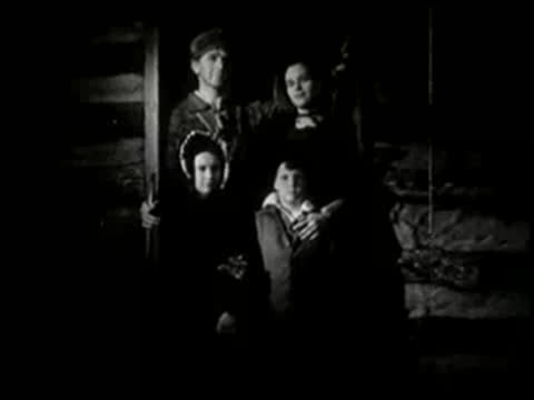 A pioneer family standing in a doorway at night, looking out.