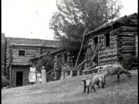 Log cabin settlement exterior with livestock and figures walking outside.
