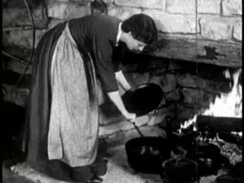 Woman cooking food in iron skillets over a hearth fire.