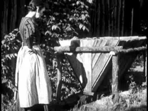 a woman working at a wooden trough or structure outdoors near a building