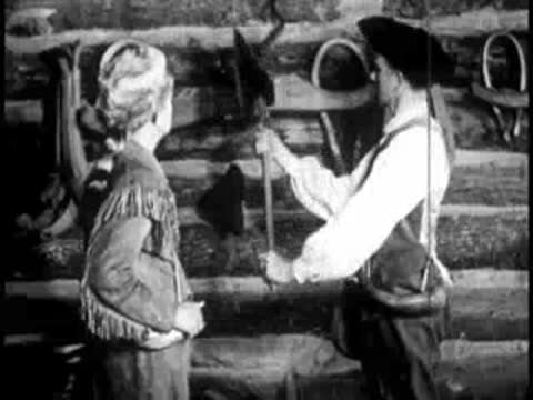 a woman handing an axe to a man wearing a hat and fringed jacket