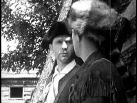 two men in historical attire talking outdoors near a wooden structure