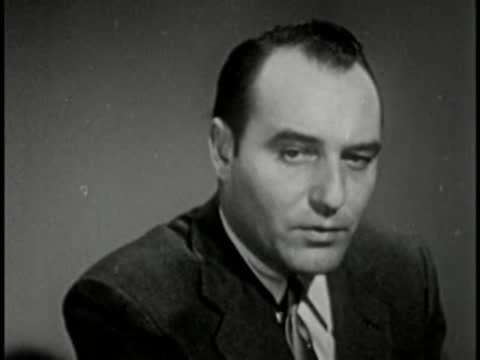 Man in dark suit speaking, vintage television studio interview setting.