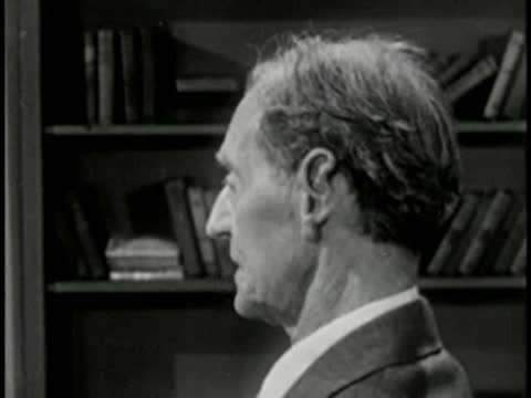 Elderly man in profile with bookshelf background, vintage television interview setting.