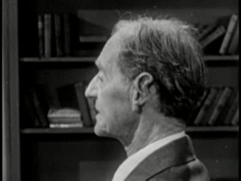 Elderly man in profile with bookshelf background, vintage television studio interview setting.