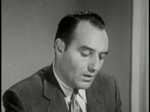 Man in dark suit looking down, vintage television studio interview setting.