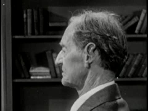 Elderly man in profile with bookshelf background, vintage television studio interview setting.