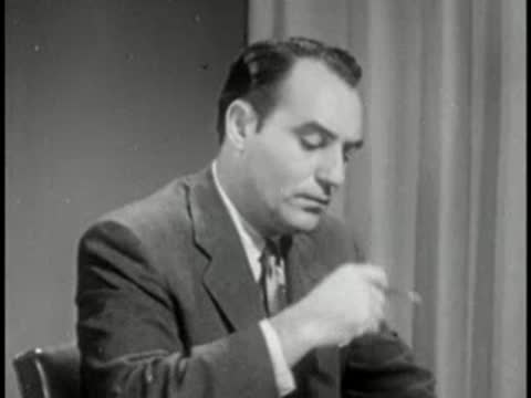 Man in dark suit touching chin and looking down, vintage television interview setting.