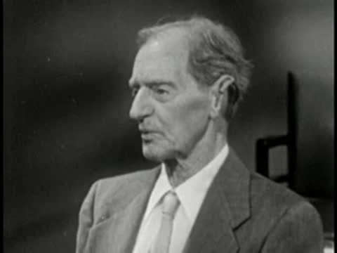 Elderly man in suit touching ear, vintage television studio interview setting.
