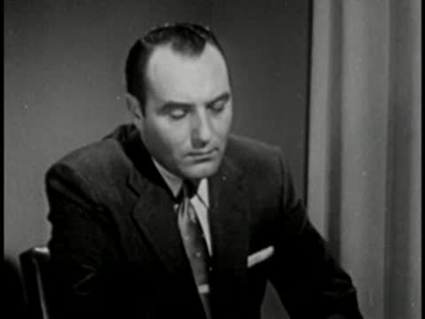 Man in dark suit looking down, vintage television studio interview setting.