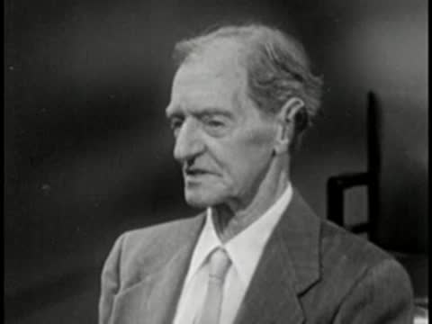 Elderly man in suit looking down, vintage television studio interview setting.