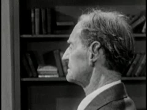 Elderly man in profile with bookshelf background, vintage television studio interview setting.