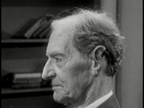Elderly man in profile with bookshelf background, vintage television interview setting.