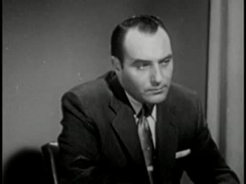 Man in dark suit writing at desk, vintage television studio interview setting.