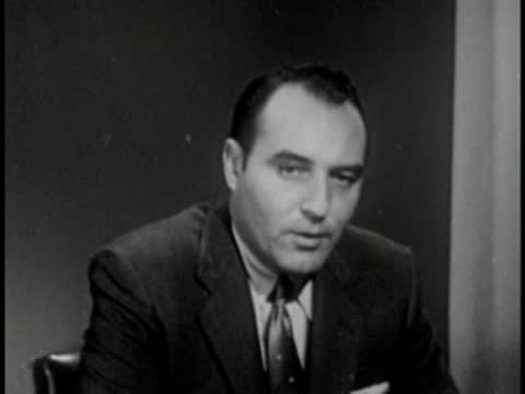 Man in dark suit speaking, vintage television studio interview setting.