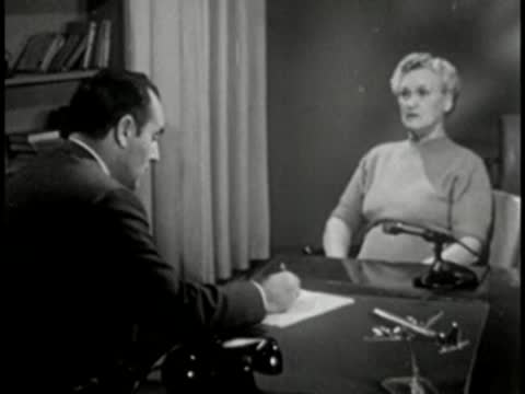 Man and woman sitting at desk, vintage television studio interview setting.