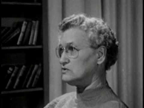 Older woman wearing glasses speaking, interview setting, vintage broadcast.