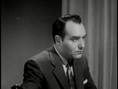 Man in dark suit speaking, vintage television studio interview setting.