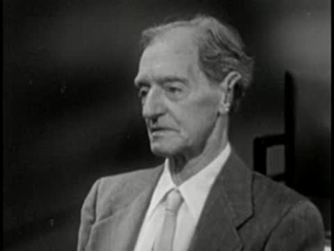 Elderly man in suit speaking, vintage television studio interview setting.
