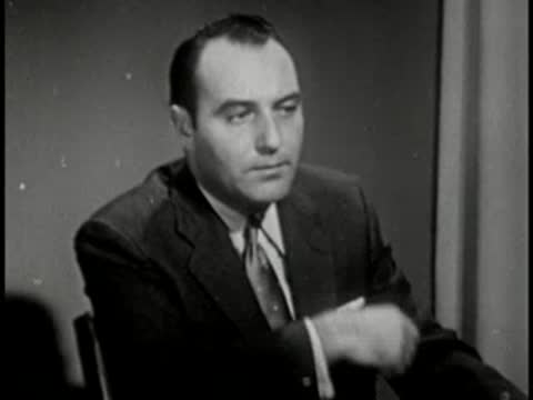 Man in dark suit touching chin, vintage television studio interview setting.