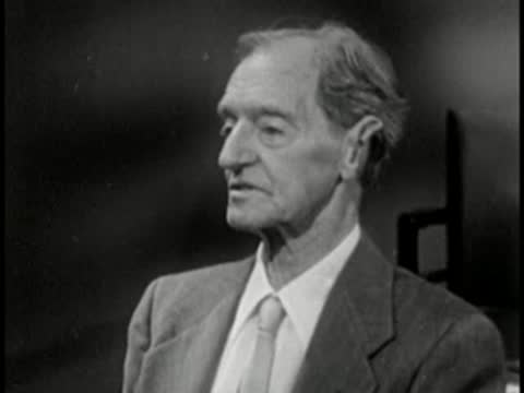 Elderly man in suit speaking, vintage television studio interview setting.