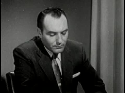 Man in dark suit looking down, vintage television studio interview setting.