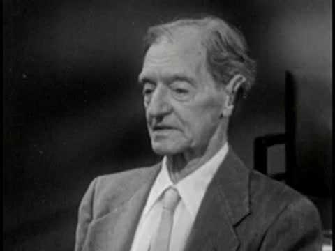 Elderly man in suit looking down, vintage television studio interview setting.