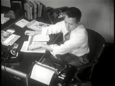 Man sitting at a large office desk, reviewing papers and typing on typewriter