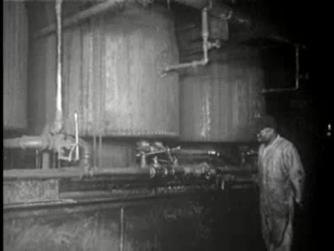 Worker stands near large industrial tanks and pipes in a factory setting.