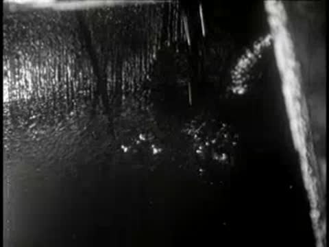 Dark fluid streams down into a large vat from overhead pipes.