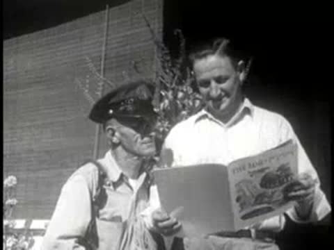 Mail carrier and a man standing outdoors and looking at a magazine together