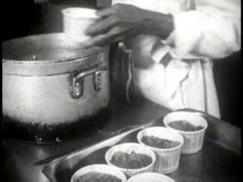 A chef in a restaurant kitchen scooping food from a large pot into containers.