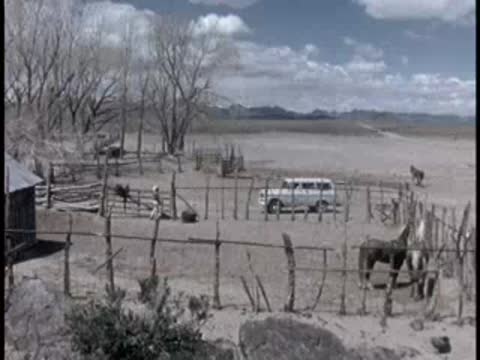 A wide shot of a dusty corral with wooden fences and distant hills under a cloudy sky