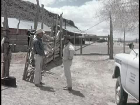 Two people conversing outdoors near a truck parked by a rustic wooden structure