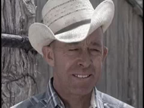 A smiling person wearing a white cowboy hat outdoors near a wooden wall