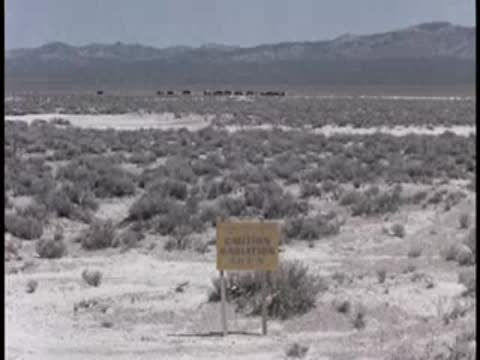 An expansive desert landscape with sparse vegetation, distant hills, and a warning sign