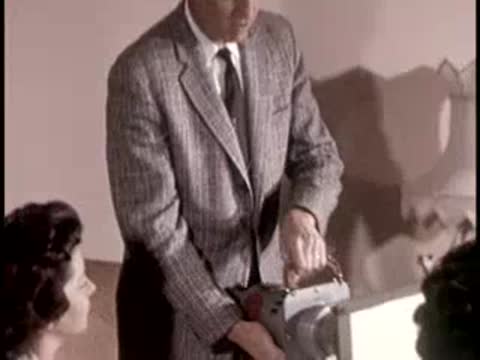 A man demonstrates a portable device to women seated indoors, retro equipment