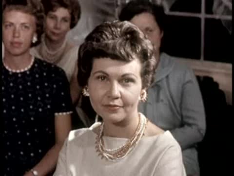 A woman smiles toward the camera in a seated audience, mid-century setting