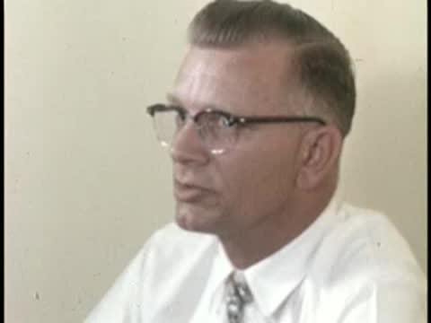 A man wearing glasses speaks while seated indoors, mid-century office environment